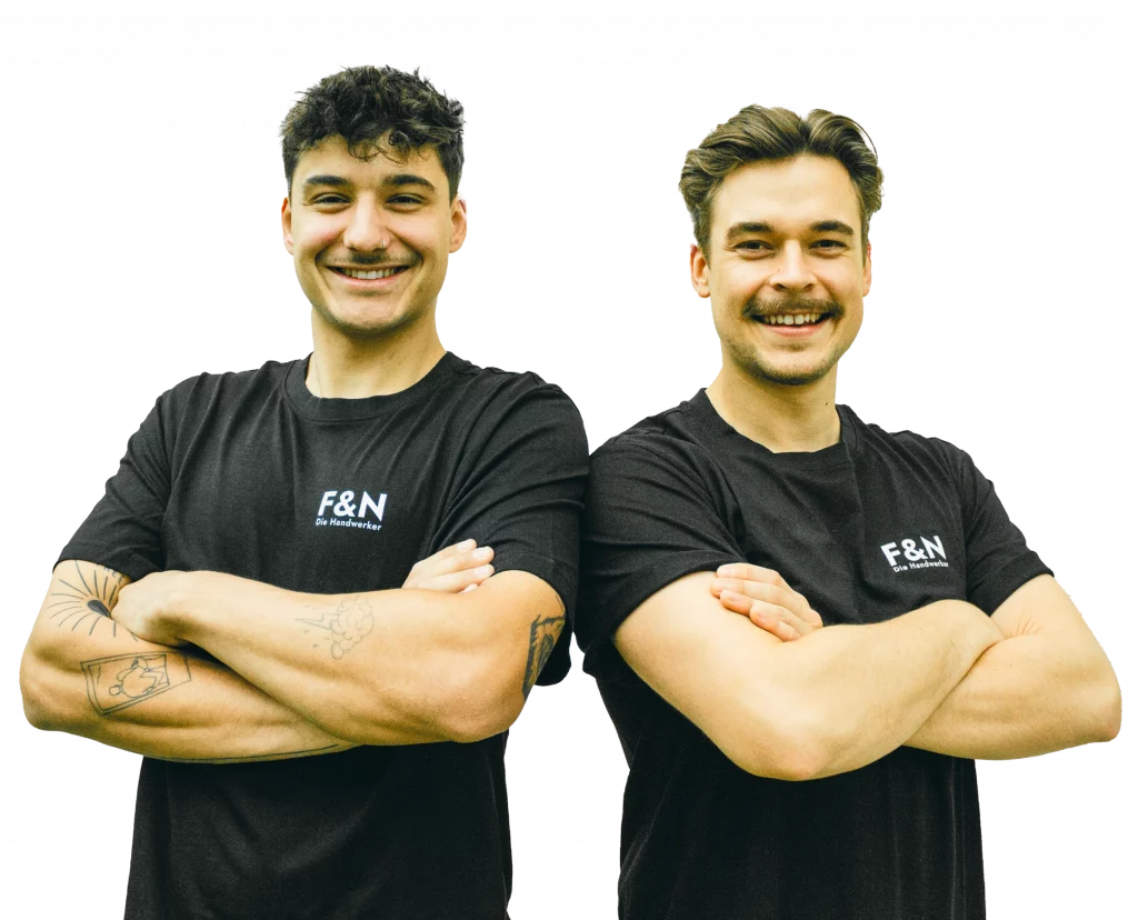 Dei Geschäftsführer Fabian und Niko stehen nebeneinander mit verschränkten Armen und lächeln selbstbewusst in die Kamera. Beide tragen schwarze T-Shirts mit dem Logo ‚F&N Die Handwerker‘. Der linke Mann hat lockiges dunkles Haar, sichtbare Tattoos auf den Armen und einen Schnurrbart. Der rechte Mann hat glattes, zurückgekämmtes Haar und ebenfalls einen Schnurrbart.