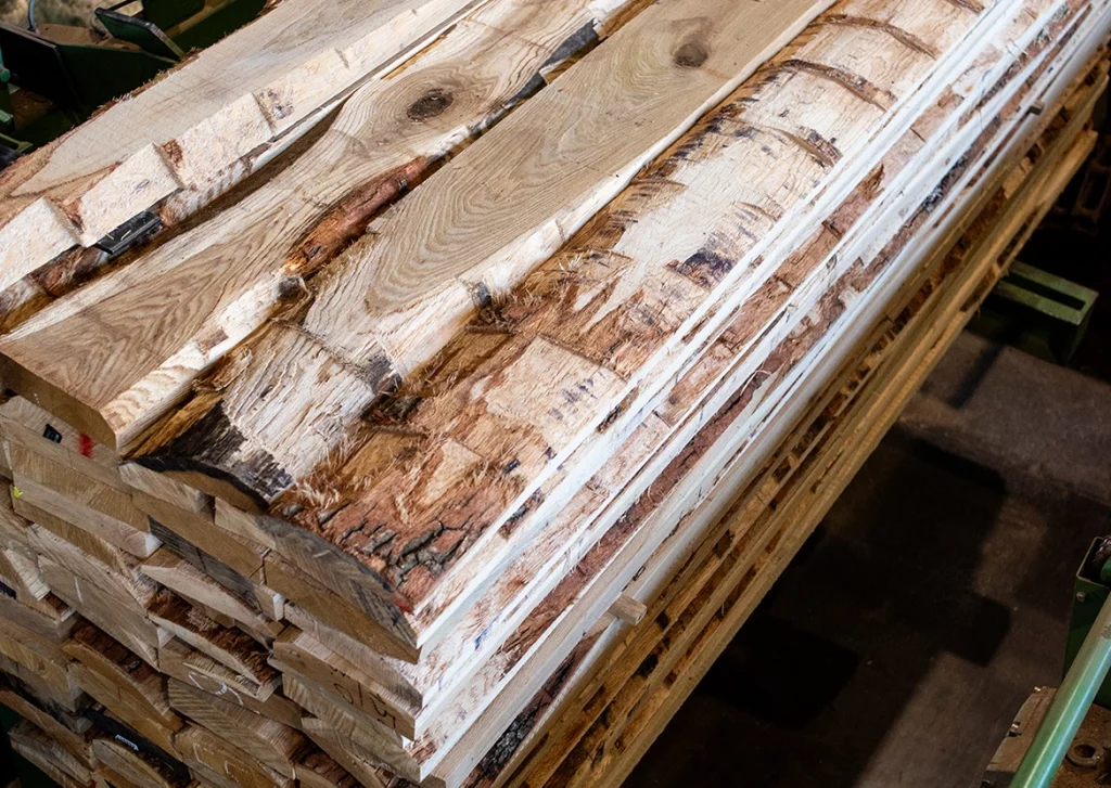 Ein Stapel grob gesägter Holzbohlen liegt in einer Werkstatt. Die Bohlen bestehen aus hellem und dunklem Holz mit natürlicher Rindenstruktur an den Kanten. Teilweise sind noch Rindenreste sowie ungleichmäßige Schnittränder zu sehen. Der Stapel liegt auf einer Holzkonstruktion, während im Hintergrund metallene Maschinenstrukturen erkennbar sind.
