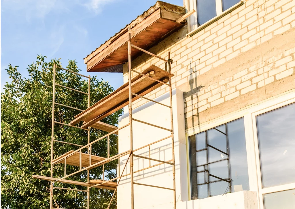 Eine Hausfassade aus hellen Ziegelsteinen mit sichtbaren Betonschichten befindet sich im Umbau. Ein Metallgerüst mit mehreren Holzplanken steht an der Wand, um Arbeiten an der Dämmung und dem Balkon zu ermöglichen. Teile der Fassade sind bereits mit weißem Dämmmaterial versehen. Die Fenster haben große Glasflächen.