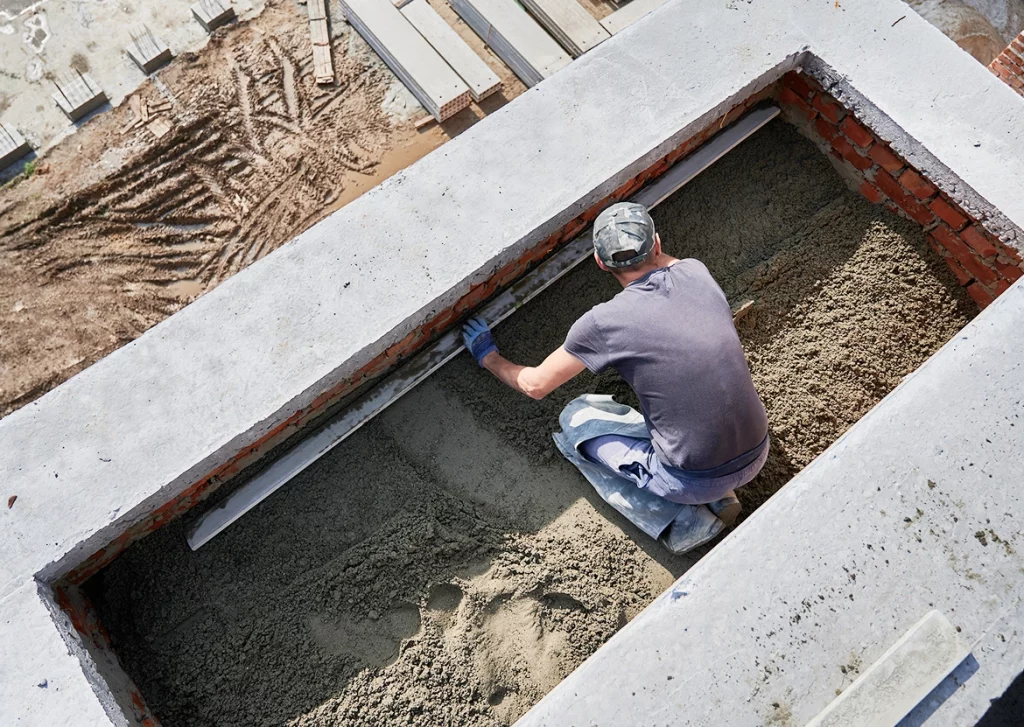 Ein Bauarbeiter in dunkler Kleidung und Camouflage-Kappe arbeitet kniend an einer Baustelle. Er glättet mit einer Wasserwaage eine Sandschicht in einer rechteckigen, von Beton und Ziegelsteinen umrandeten Öffnung. Er trägt blaue Handschuhe und hat eine Schutzmatte unter seinen Knien. Im Hintergrund sind Baustellenmaterialien wie Betonplatten und aufgewühlte Erde mit tiefen Reifenspuren sichtbar.