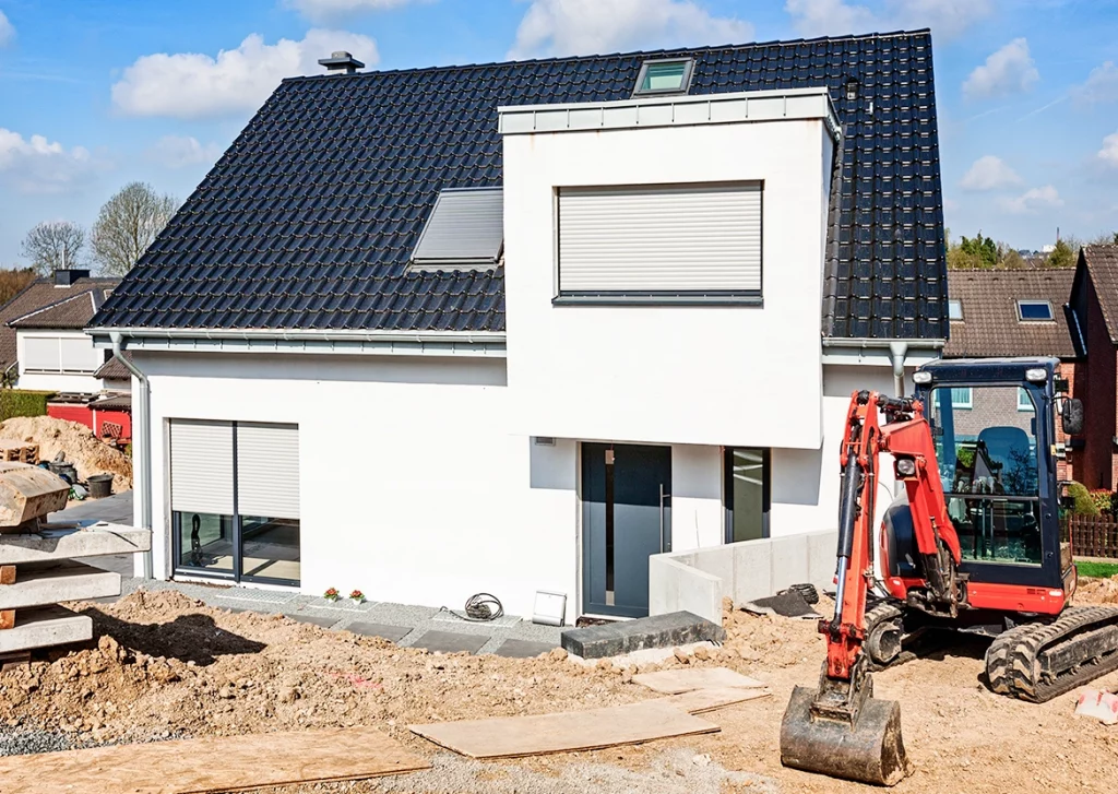 Ein modernes, weißes Einfamilienhaus mit dunklem Ziegeldach steht in einer Neubausiedlung. Die Rollläden der Fenster sind größtenteils geschlossen. Vor dem Haus befindet sich eine Baustelle mit aufgeschütteter Erde und Bauplatten als provisorischem Weg. Ein kleiner roter Bagger mit ausgefahrenem Arm steht im Vordergrund. Neben dem Eingang sind graue Steinplatten als zukünftiger Gehweg verlegt.
