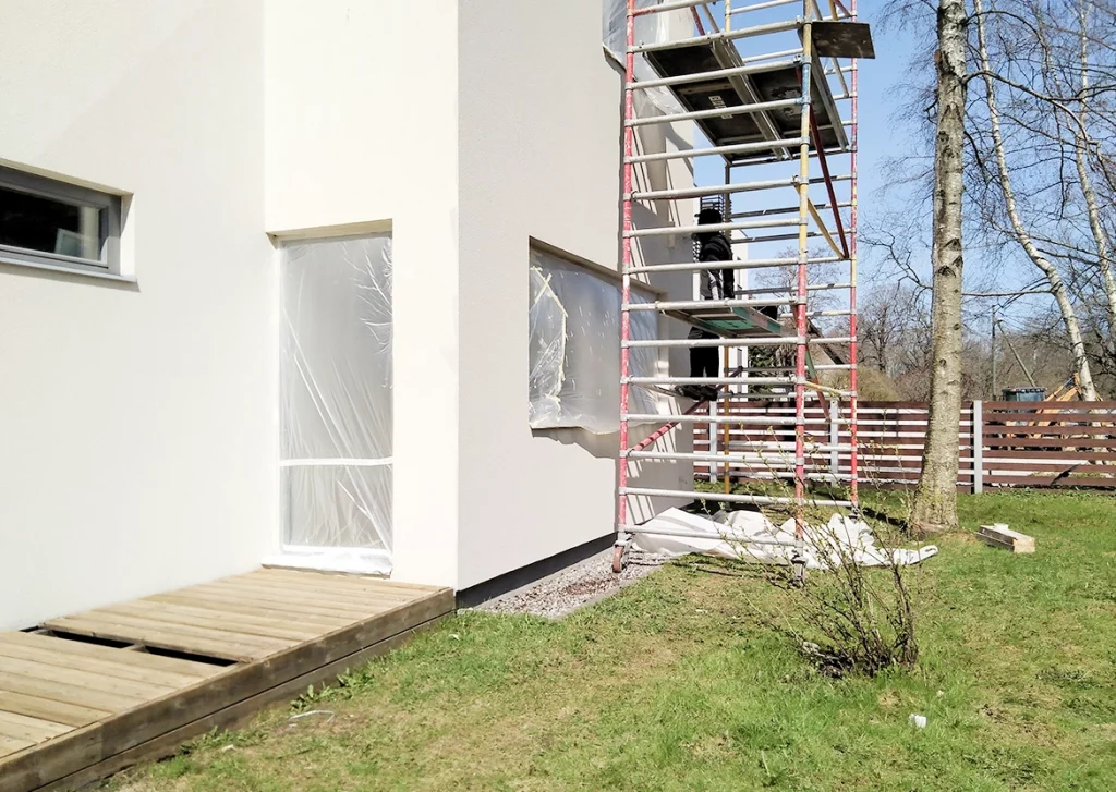 Ein modernes, weißes Wohnhaus wird renoviert. Ein Gerüst mit mehreren Plattformen steht an der Fassade, und einige Fenster sind mit transparenter Plastikfolie abgeklebt, um sie zu schützen. Ein hölzerner Gehweg führt zur Eingangstür. Im Vordergrund ist eine Rasenfläche mit einem kleinen Strauch zu sehen.